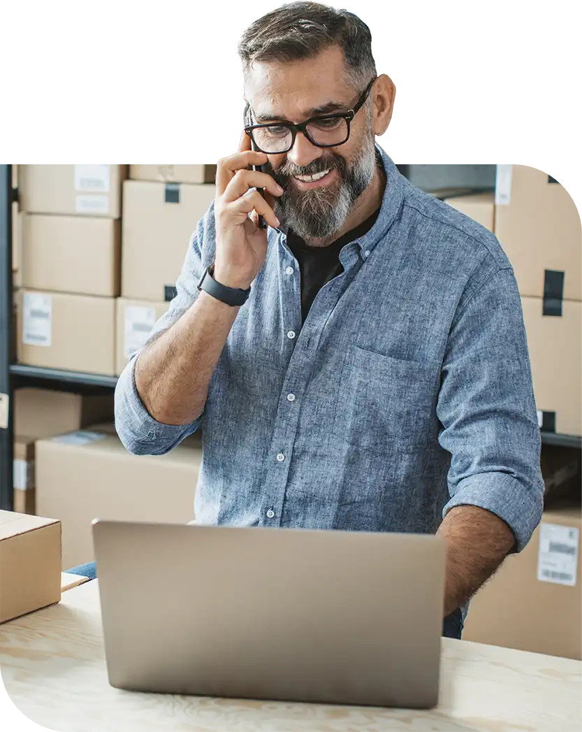 Small business owner on the phone while working on a laptop in a warehouse full of shipping boxes