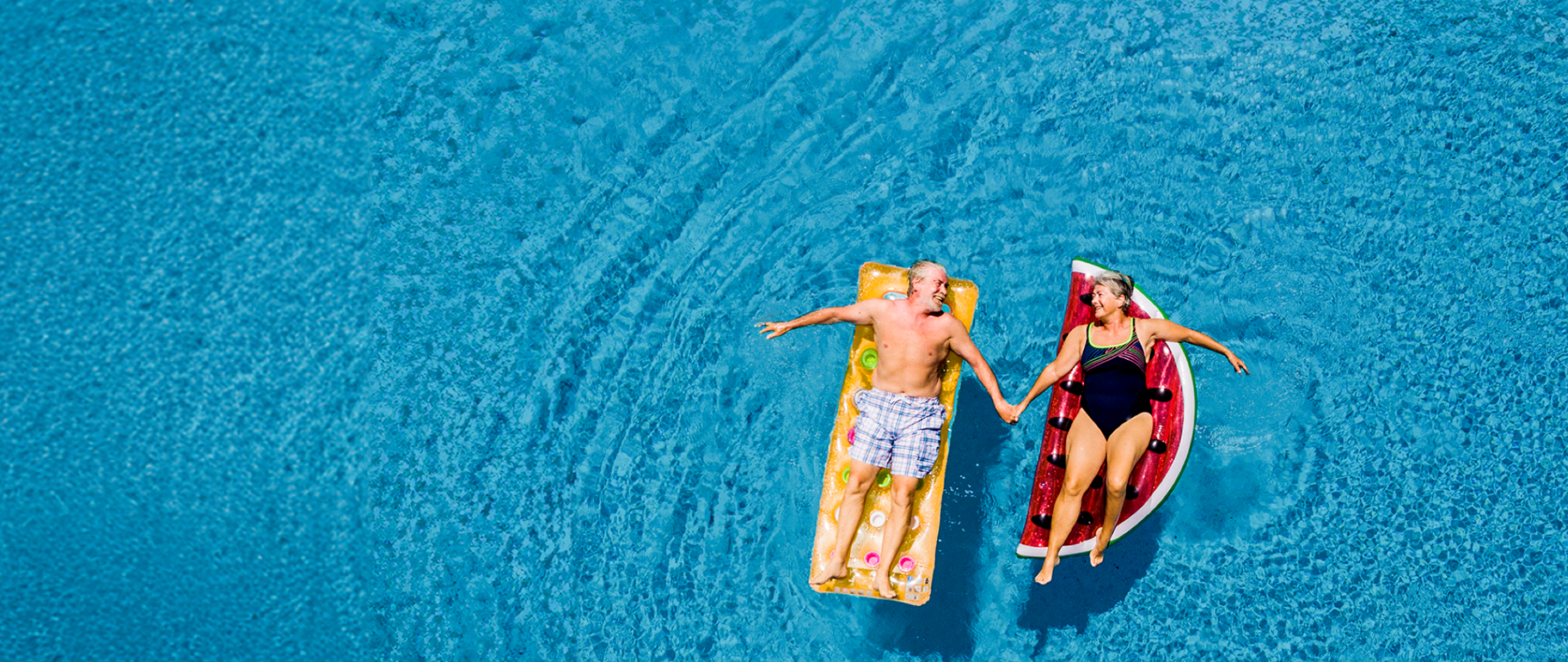 bird's eye view photo of retired couple 65+ floating in a pool on vacation with a watermelon floatie holding hands on a sunny day.